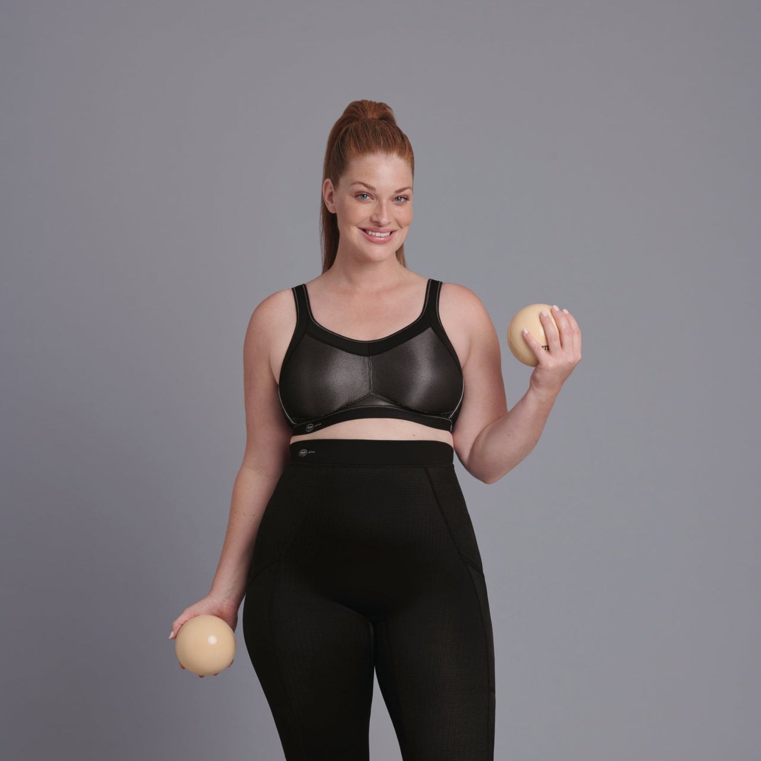 Vrouw in zwart-grijze glanzende sportbeha met een bal in elke hand, studiosetting