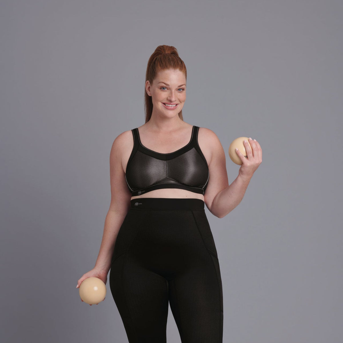 Vrouw in zwart-grijze glanzende sportbeha met een bal in elke hand, studiosetting